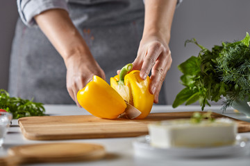 Woman's hands cut yellow pepper on a wooden board on the kitchen table. Cooking salad