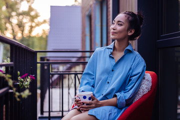 From the balcony. Joyful Asian woman looking at the street while holding a cup of tea