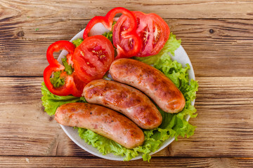 Ceramic plate with grilled sausages, sliced tomatoes and lettuce leaves on wooden table. Top view