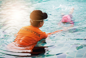 Female instructor teaches kid how to swim. Kid with goggles in water swimming to the trainer. Happy kid in modern swimming pool. Concept of fun, leisure and recreation.
