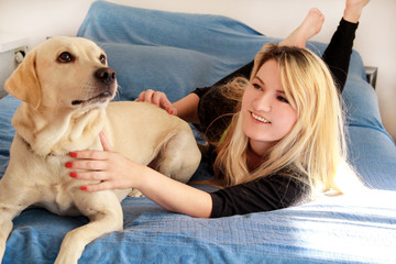 Woman with her dog in bed at home, relaxing in bedroom. Beautiful girl is playing, together and petting with dog in bed. Yellow labrador retriever climbed into bed with her owner. Lovely dog, pet.