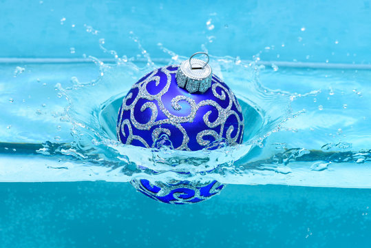 Holidays and vacation concept. Festive decoration for Christmas tree, blue ball dropped into water with splashes, blue background. Christmas decoration or toy for Christmas tree swim in pool