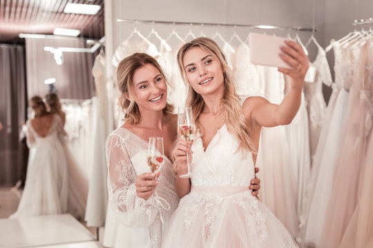 Beautiful photo. Joyful positive brides holding glasses with sparkling wine while taking a selfie together