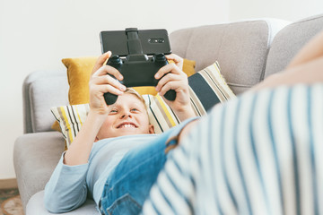 Teenager boy lying on the cozy sofa and  enthusiastically plays game using the gamepad with...