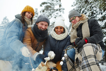 Positive excited young multi-ethnic friends in winter coats sitting by campfire and roasting marshmallows on fire at camping in forest