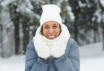Obraz premium Cheerful excited attractive young woman in mittens wearing white hat and scarf looking at camera and standing under snow in winter forest