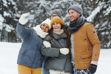 Group of cheerful excited young friends in puffy jackets laughing while taking selfie on smartphone in winter forest