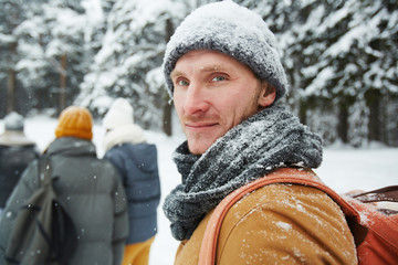 Content handsome young male hiker in snowy hat and scarf turning back while looking at camera in winter forest, he following group of travelers