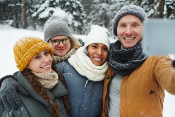 Positive excited young friends in warm hats and scarves posing for selfie and embracing in winter park