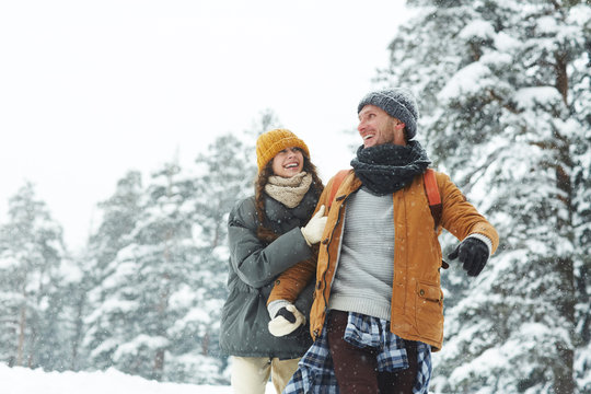 Jolly beautiful young couple in puffy jackets holding hands and laughing while enjoying winter stroll, hiking in forest - Powered by Adobe