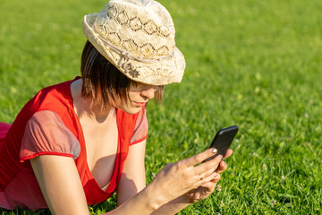 attractive girl in red dress is lying on a green grass and palying with her phone