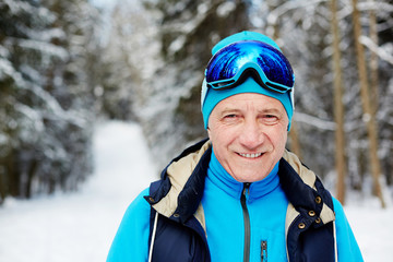 Active senior man in sportswear and skiing goggles looking at you during training in winter forest