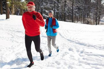 Happy mature man and woman in sportswear running down snowdrift on winter morning