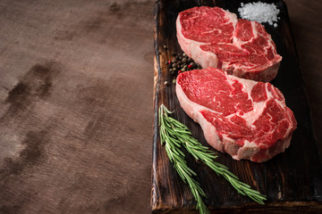 Two fresh raw rib-eye steak on wooden Board on wooden background with salt, pepper and rosmary in a...