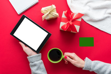 Obraz premium Female hands, woman holding a tablet pc and Cup of coffee or tea. Business card, red table, gifts boxes for the holidays, background with copy space for advertisement, top view