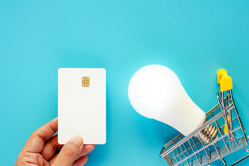 Hand holding a blank white credit card with glowing LED light bulb and mini shopping cart or trolley for energy savings and planning concept