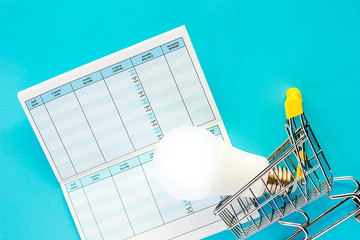 Glowing LED light bulb with mini shopping cart or trolley and savings book on blue background for energy savings and planning concept