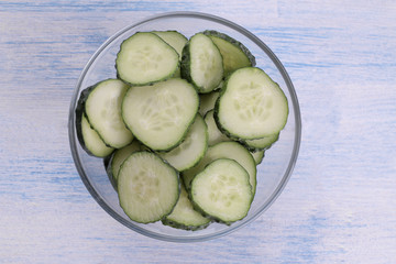 Slices of fresh cucumber in a glass bowl on a blue wooden table. view from above