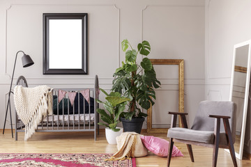 Mockup poster in black frame above grey wooden crib in stylish baby room interior with green plants...