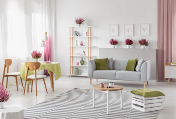 Living room interior with dining table in real photo with window with curtains, sofa with cushions, rack with books and decor and heathers