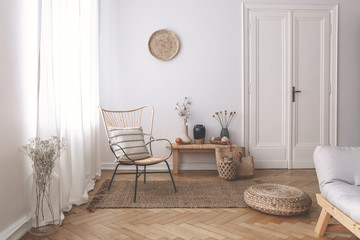 Sheer white curtains on the window of a white living room interior with a striped, linen pillow on a modern wicker chair