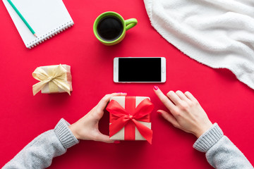 Female hands, a woman holding a gift and uses smart phone. Red table, white smart phone, gifts boxes for the holidays, background with copy space for advertisement, top view