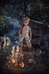 Naklejka na ściany i meble Caveman, manly boy at the fire. Scary young primitive boy outdoors near bonfire. Witch craft concept. Angry caveman, manly boy with horns near bonfire. Prehistoric tribal man outdoors on nature