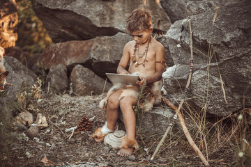Fototapeta na wymiar Caveman, manly boy using tablet PC. Funny young primitive boy outdoors. Evolution degradation concept. Calm boy outside against rocky background. Prehistoric tribal man outside on wild nature with