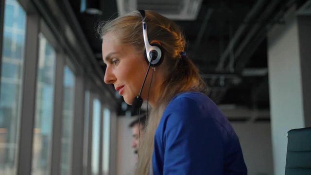 Close-up of young businesswoman talking on headset with perfect smile. Attractive businesspeople speaking with customers at background.