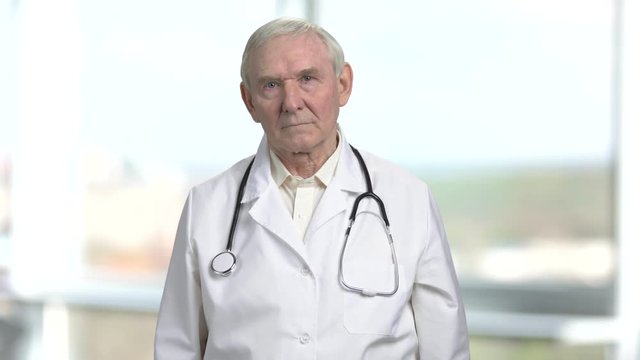 Old doctor get rid of excessive sweating. Portrait of frowning senior doctor with stethoscope. Bright windows background.