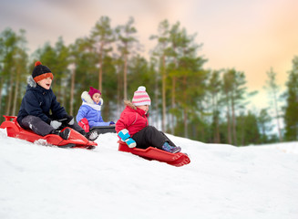 Fototapeta na wymiar childhood, sledging and season concept - group of happy little kids sliding on sleds down snow hill over winter forest background and snow