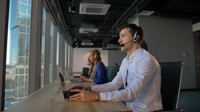 Attractive business person waring white formal shirt in office call center talking with client and then smile to camera. Concept of successful and cheerful solving of customer problems online.