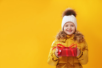 Happy little girl preschooler in a winter jacket and  in a knitted hat is holding a box with a gift. The concept of giving and receiving gifts. Yellow on a yellow background, red.
