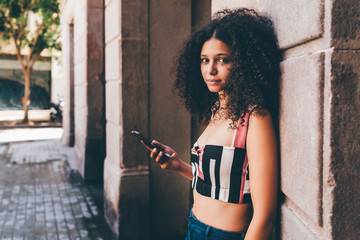 Portrait of a young beautiful Moroccan female tourist leaning against the wall outdoors and holding her smartphone; charming curly African-American woman with the cell phone on the street of Barcelona