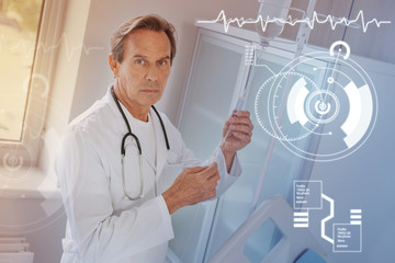 Serious medical worker. Attentive experienced doctor looking concentrated while standing with a stethoscope on his neck and preparing an infusion for his patient