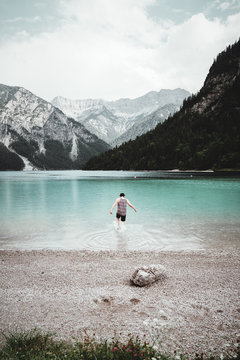 junger mann mit Hut und tank-top rennt in das wasser des Plansees im hintergund die Alpen