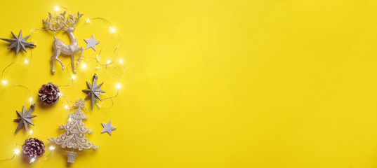 Christmas and new year concept. Greeting banner with copyspace. Shiny silver deer, stars, fir-tree, garland, bokeh on yellow background. Top view, flat lay - obrazy, fototapety, plakaty