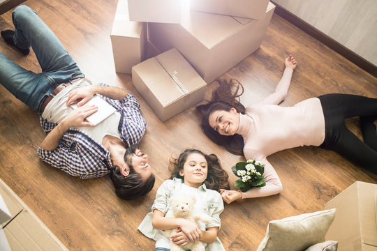 Family portrait where everybody is lying on the floor. Small girl has her teddy bear in the hnds. There is a plant near young woman's hand. Young man is holding a tablet or notebook.