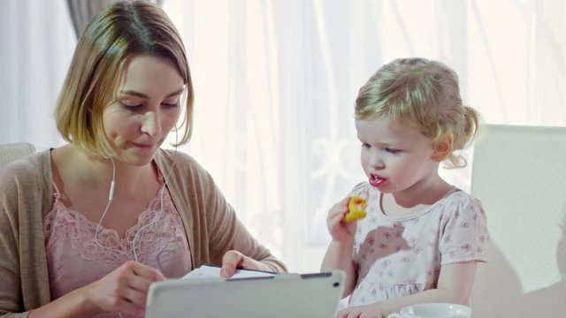 PAN of busy young mother sitting at dining table with cute toddler girl eating croissant and talking to her while writing notes in planner