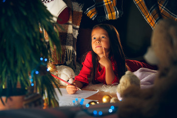 Merry Christmas and Happy Holidays. Cute little child girl writes the letter to Santa Claus near Christmas tree at home indoor. The holiday, childhood, winter, celebration concept