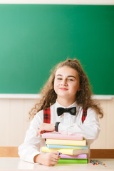 schoolgirl in school uniform sitting at her desk with books and pencils on the background of a green board