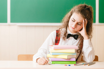 schoolgirl in school uniform sitting at her desk with books and pencils on the background of a green board