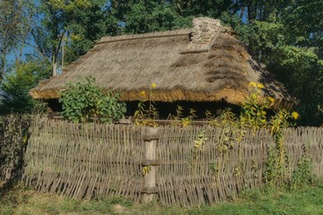 an old cottage with a straw roof surrounded by an old wicker fence with a garden with sunflowers