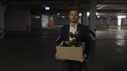 Very Emotional disappointed Corporate worker gets fired and walking down parking lot with carton box and documents with a table flower. Almost crying.