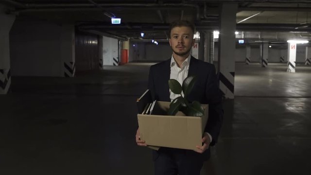 Very Emotional disappointed Corporate worker gets fired and walking down parking lot with carton box and documents with a table flower. Almost crying.