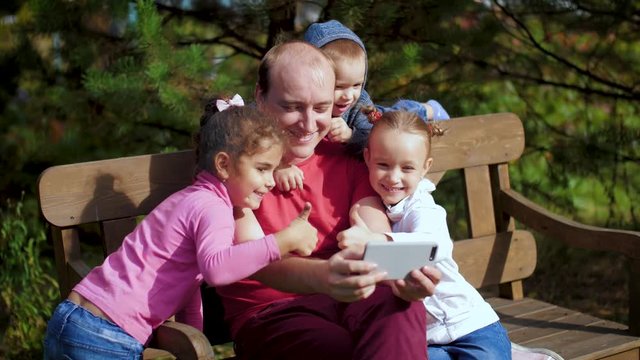 A man with children photographed on a smartphone sitting on a bench. Happy family taking selfie in the park. Family video call on mobile Internet. 4k 60fps slow motion.