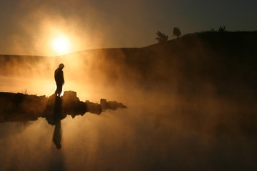 Warm sunrise silhouettes a hiker against a cold early morning mist across a lake. The silhouette of...