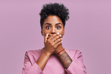 Terrified dark skinned cute young female with Afro hairstyle, covers mouth with great fear, tries not to cry, has scared shocked expression, wears striped sweater, isolated lavender background