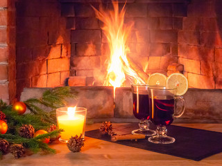 Two glasses with mulled wine, a candle, fir branches with decorations on a wooden table against the background of a burning fireplace. Romantic christmas concept