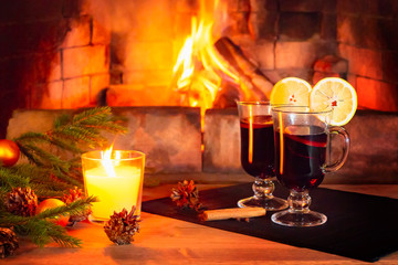 Two glasses with mulled wine, a candle, fir branches with decorations on a wooden table against the background of a burning fireplace. Romantic christmas concept